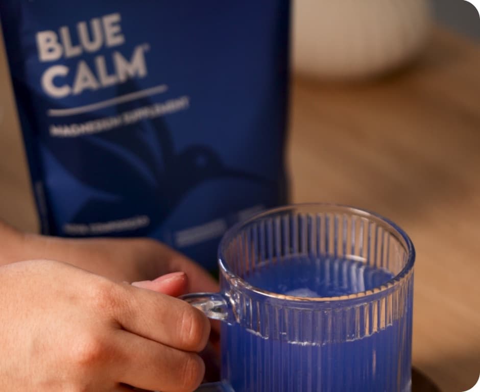 Mão segurando uma caneca de vidro com bebida azul ao lado do sachê Blue Calm