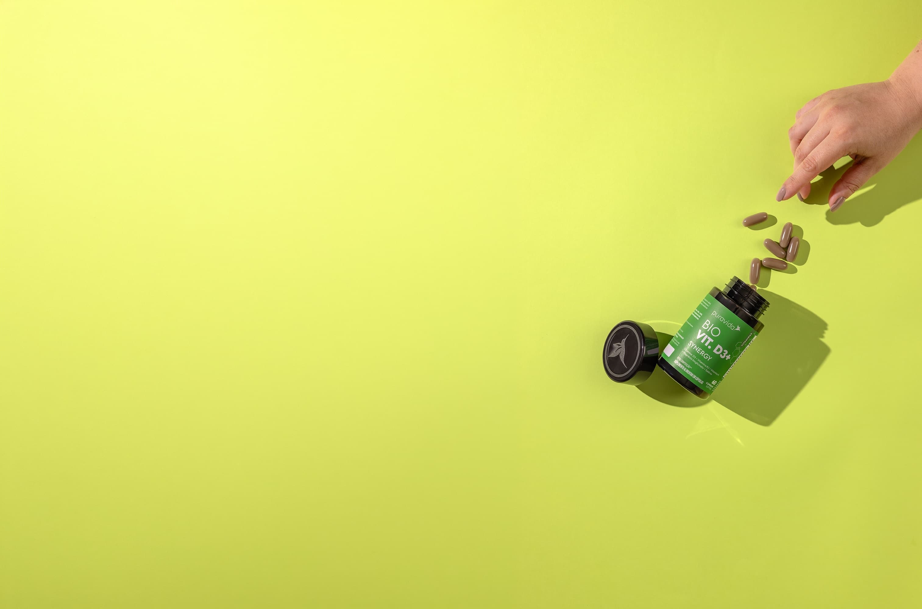 Mão segurando o frasco do Bio Vit D3+ Synergy com as cápsulas
